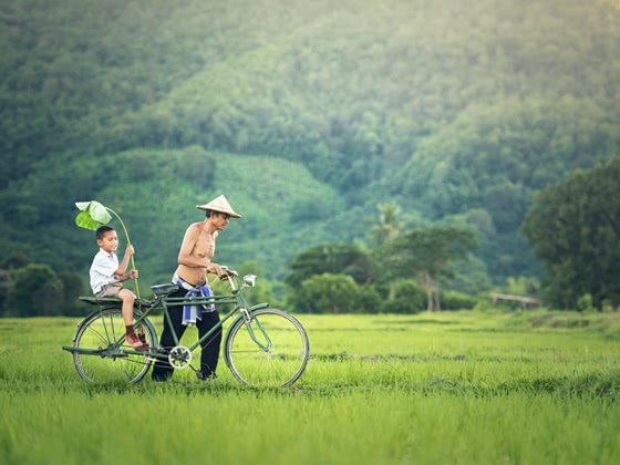 Man pushing a bike with a young boy sitting on it in a green field