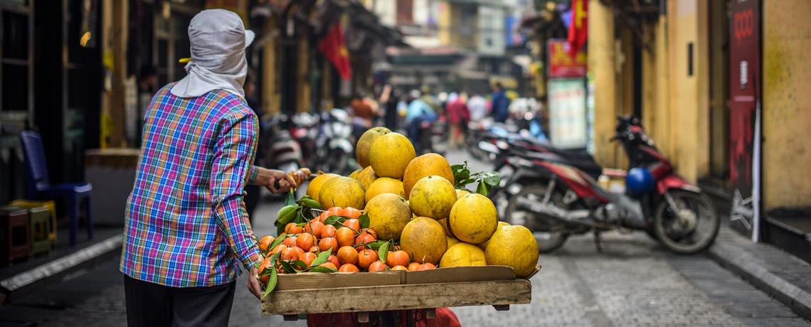 Person walking a bike laden with fruit along a narrow street