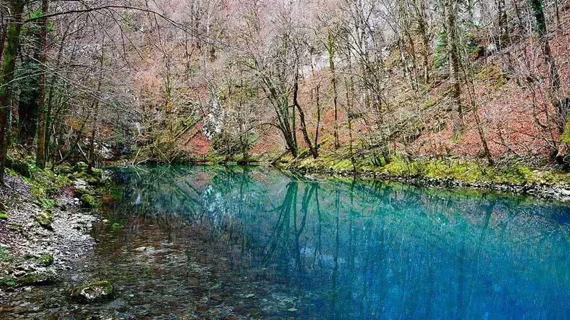 small pond in a forest with the sky and trees reflected in the turqoise water.